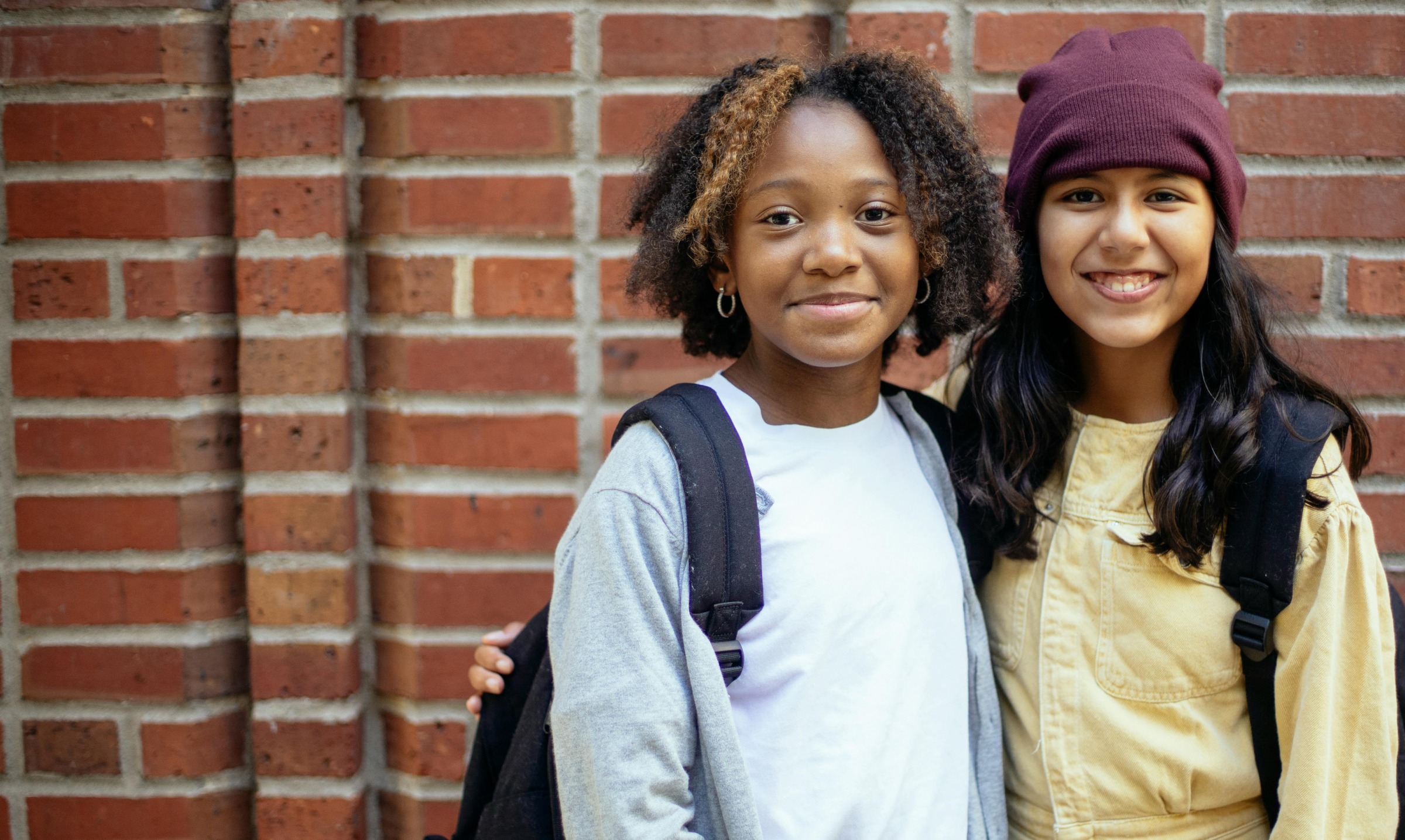 Two young teenage girls smiling while standing in front of a brick wall.