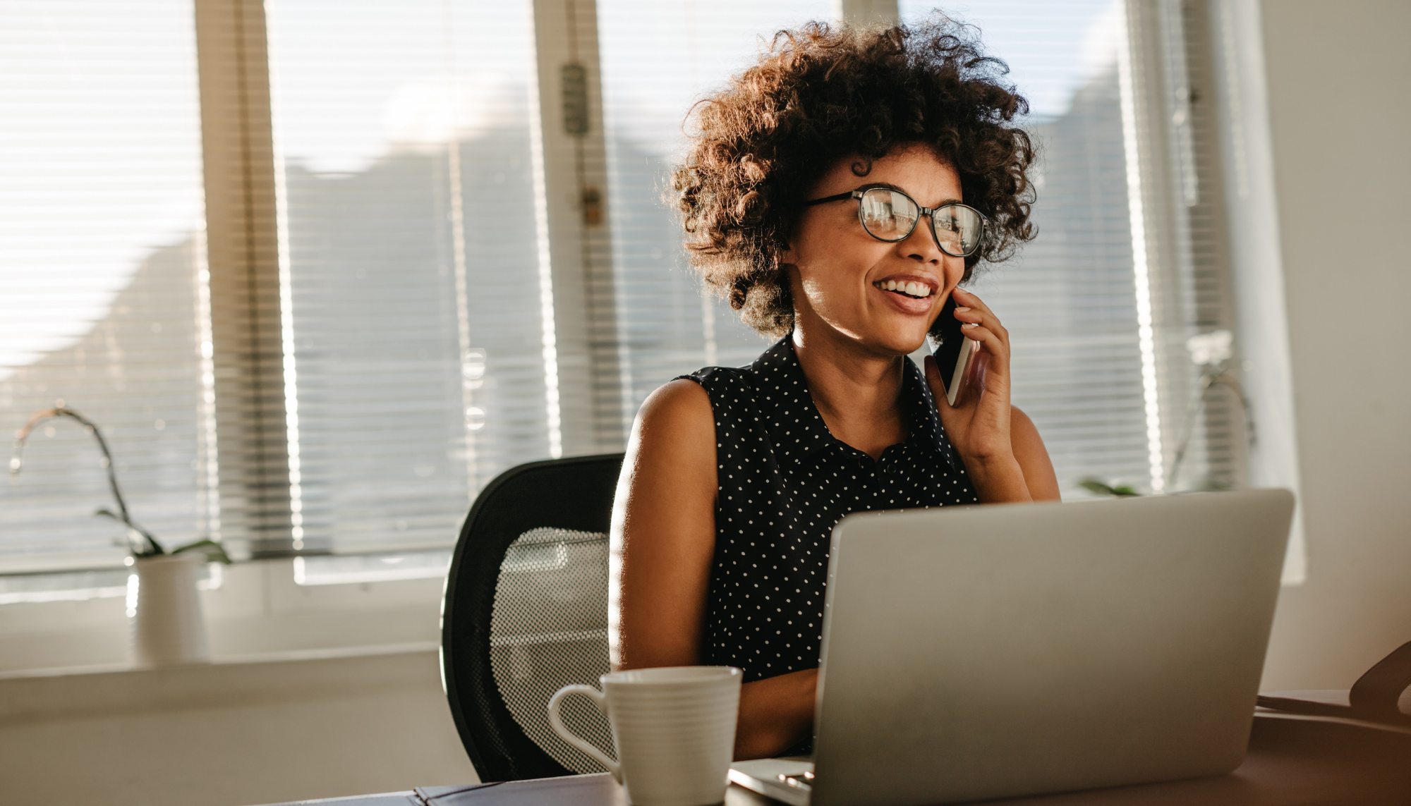 Woman smiling talking on the phone sitting in front of a laptop with a bright window behind her