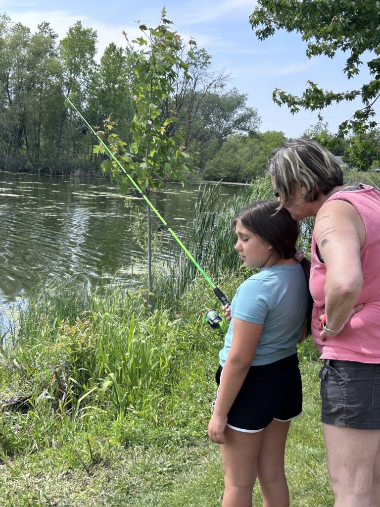 An adult and a young girl stand side by side near the edge of a pond, fishing with a bright green fishing pole. Green trees and reeds surround the water on a sunny day.