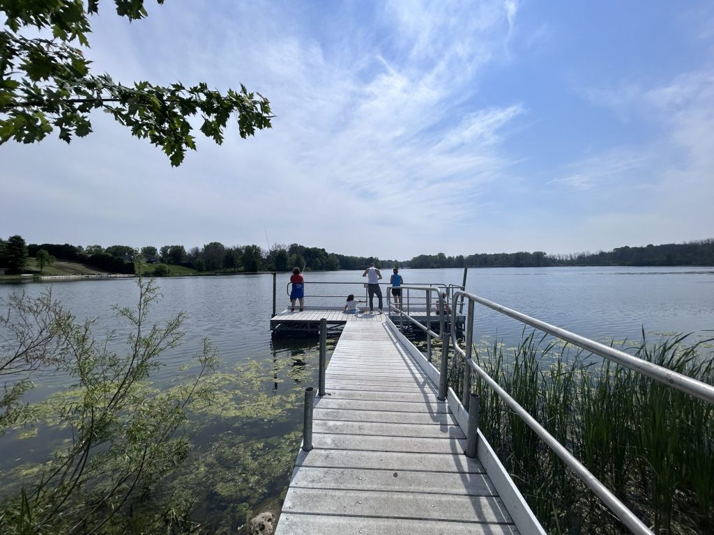 Several people fish from a metal dock extending into a large, clear lake. Green vegetation lines the shore, and a few wispy clouds stretch across the bright blue sky.