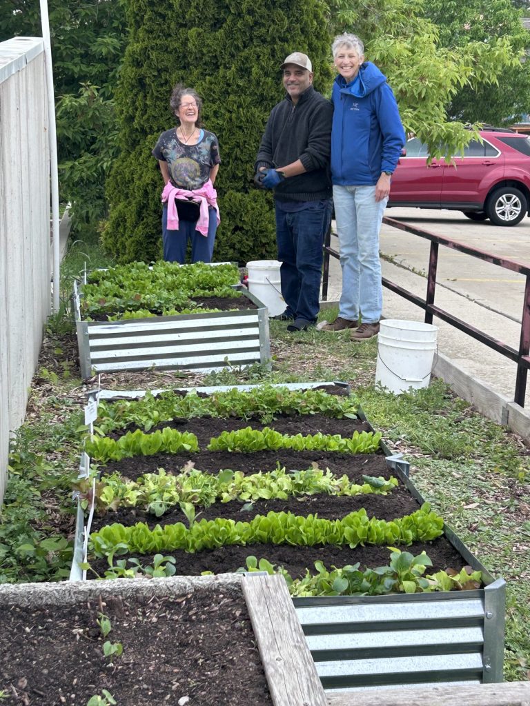 Three smiling people stand behind raised garden beds filled with rows of green leafy vegetables. The garden is next to a building and a red car is visible in the background.