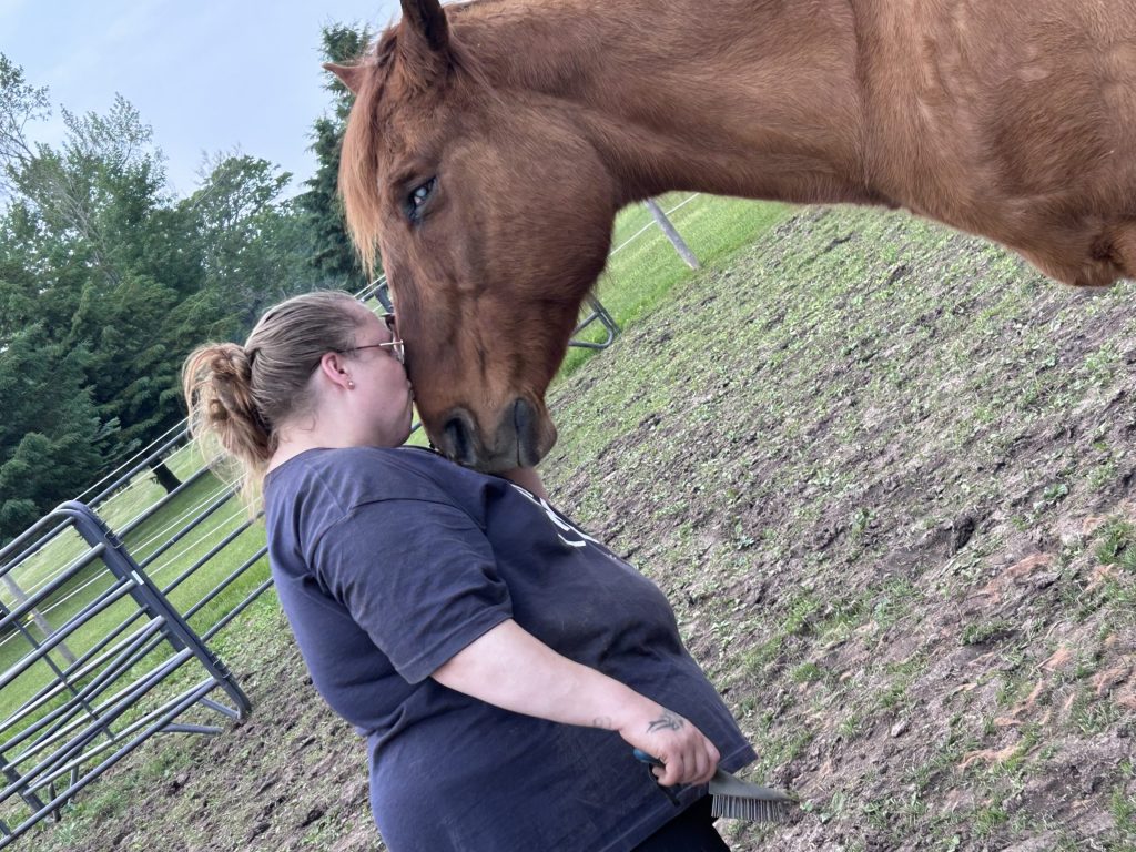 A person stands close to a brown horse, gently pressing their forehead against the horse’s face in a quiet moment of connection. The person holds a brush and stands in a grassy paddock with trees in the background.