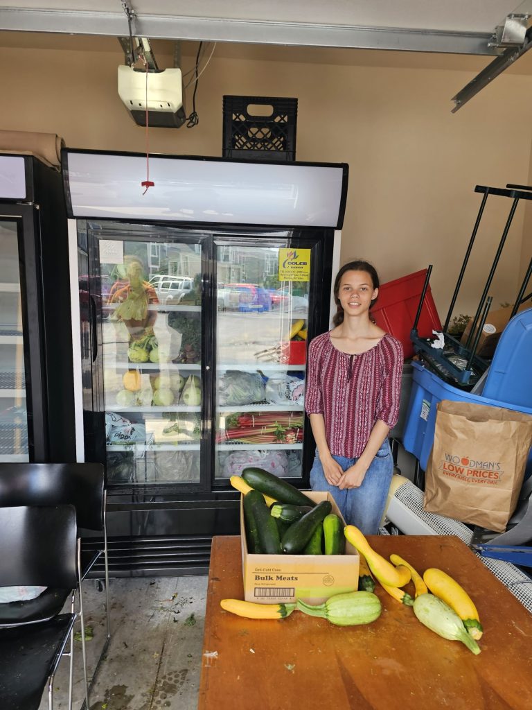 A young woman stands in front of a commercial refrigerator filled with fresh produce at a community pantry. A table in front of her holds zucchini and yellow squash.