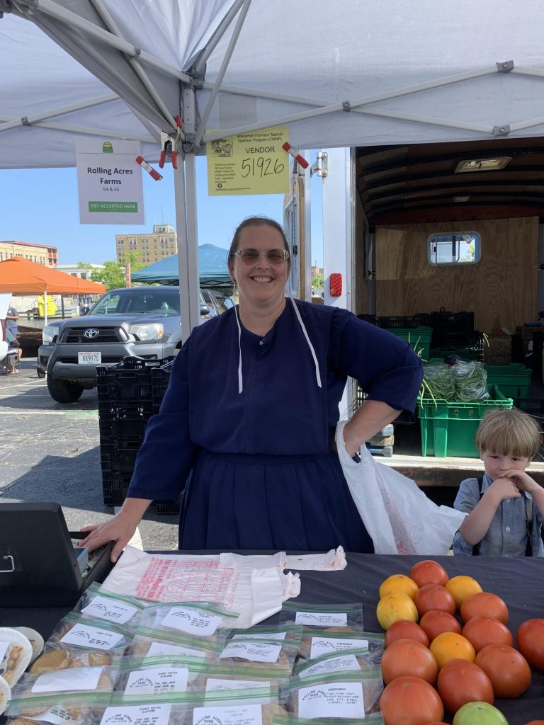 A woman wearing a navy dress and apron stands behind a farmers market table with tomatoes and packaged baked goods. A young child stands beside her. Signs for Rolling Acres Farms hang above the booth.