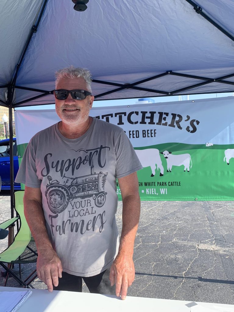 A man wearing sunglasses and a “Support Your Local Farmers” T-shirt stands behind a booth at the farmers market with a Fletcher’s Grass-Fed Beef banner displayed behind him.