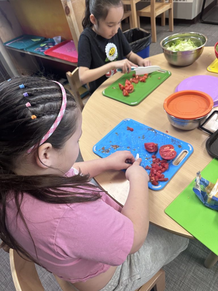 Two young children sitting at a table and chopping tomatoes.