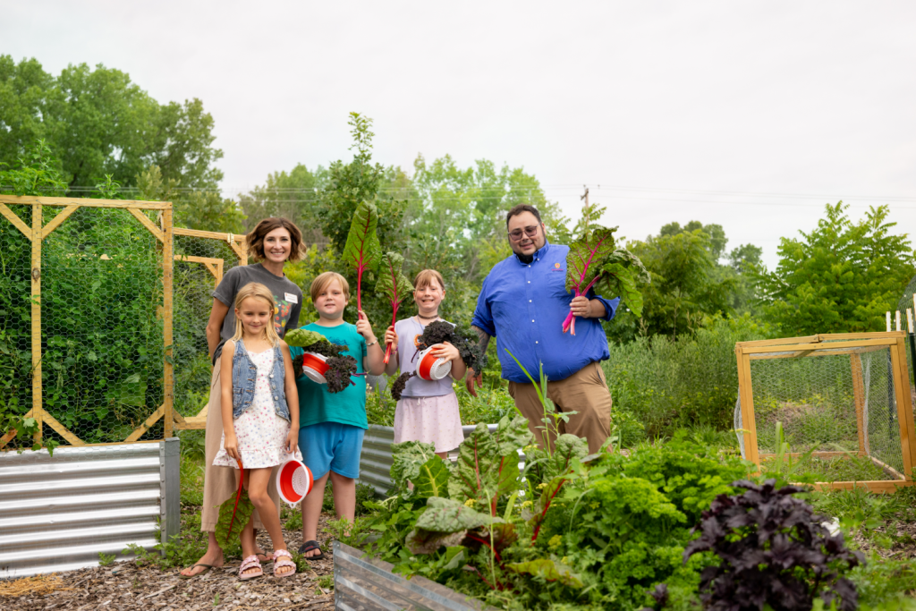 Children and adults gather around a garden, holding large leafy vegetables and looking at the camera smiling.