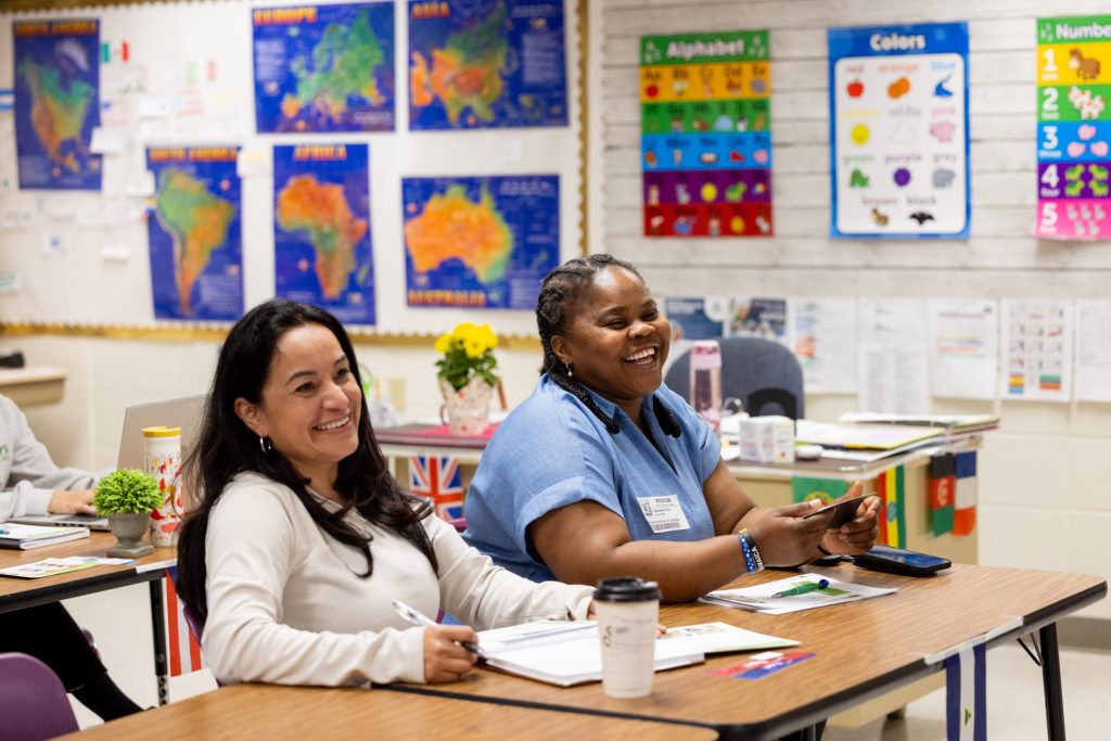 Two women sit at a desk in a classroom, smiling and laughing. The room features educational posters including world continent maps, an alphabet chart, colors chart, and numbers chart, along with small flags from various countries displayed nearby.