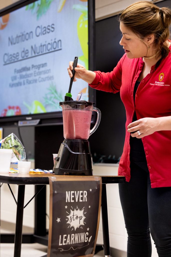 A woman in a red Extension-branded cardigan stirs a pink smoothie in a blender during a nutrition class demonstration. A screen behind her displays "Nutrition Class / Clase de Nutrición" with program details for the FoodWise Program, UW-Madison Extension, Kenosha & Racine Counties.